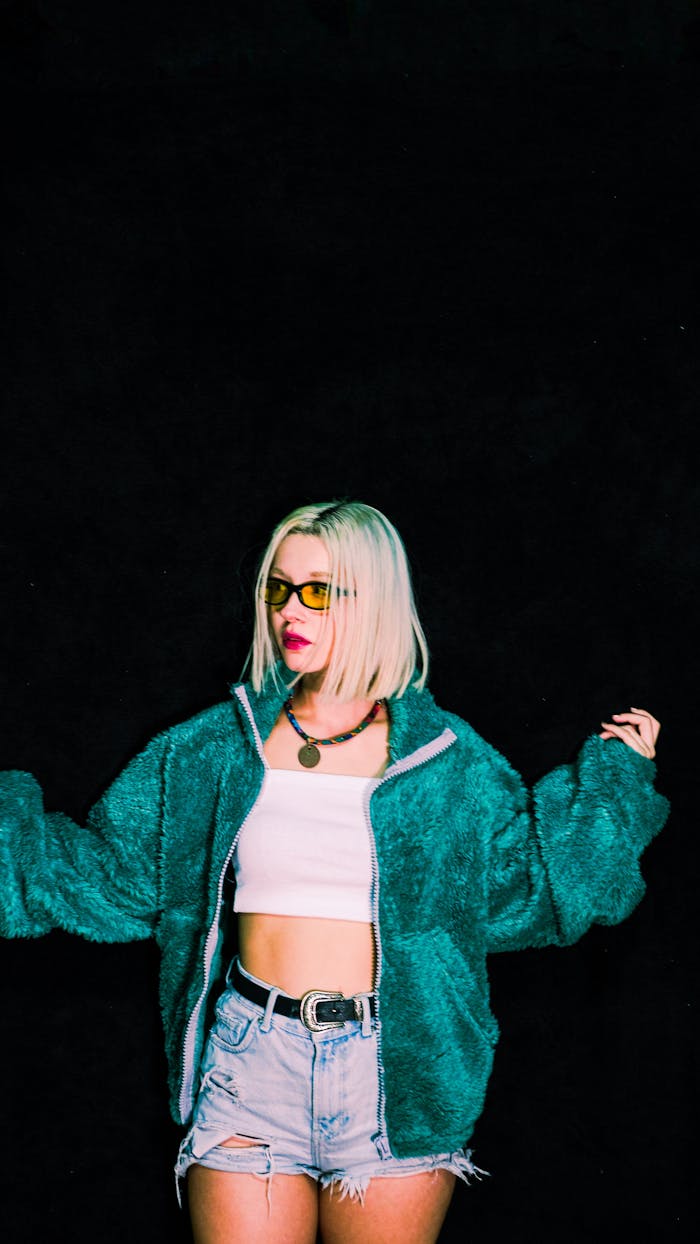 Fashionable young woman posing in a turquoise jacket and denim shorts against a dark studio backdrop.