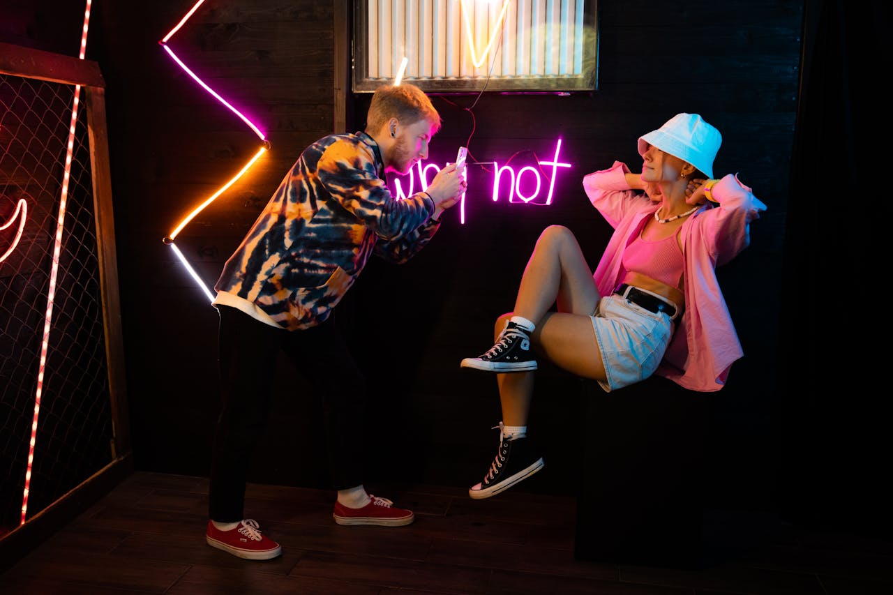 Stylish photoshoot with neon lights, featuring a man taking photos of a fashionable woman posing.
